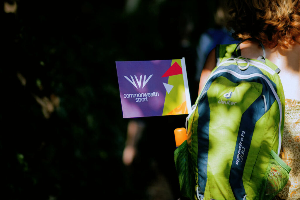 Event photography from the Commonwealth Road Race in Warwick. bright sunlight bounces off a miniature spectator flag hanging from a green backpack. Taken by Exeter photographer Dennis Cook.