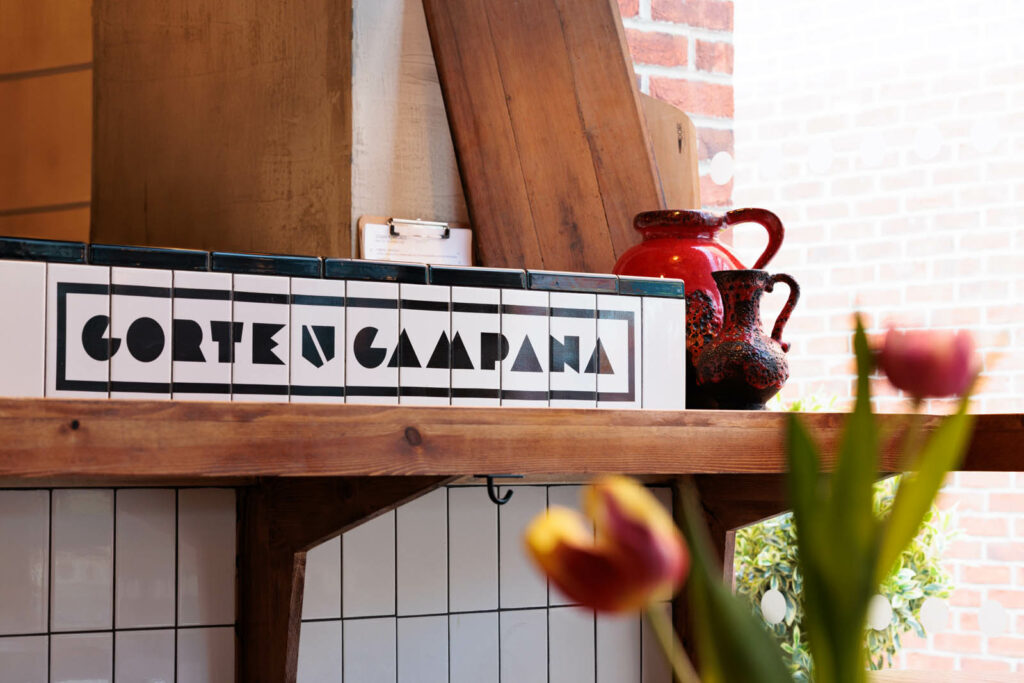 Sunlight pours through a window onto a white tiled counter top which features the Corte Campana logo. Colourful jugs and flowers surround the counter. An example of Corte Campana's brand design and visual identity in a larger, restaurant context.