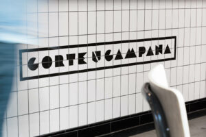 A person in blue jeans walks past a bar tiled with white metro style tiles. The tiles are decorated with the Corte Campana restaurant logo design Exeter creative Dennis Cook developed with the restaurant team.