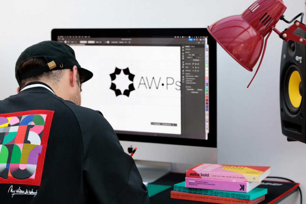 Graphic designer Dennis Cook is wearing a black jacket and cap. He works in front of his computer on a logo design project in his Exeter design agency space.
