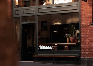 A wide shot of the Alma coffee shop in Exeter, Devon. There is a logo on the window and you can see flowers and furniture inside.