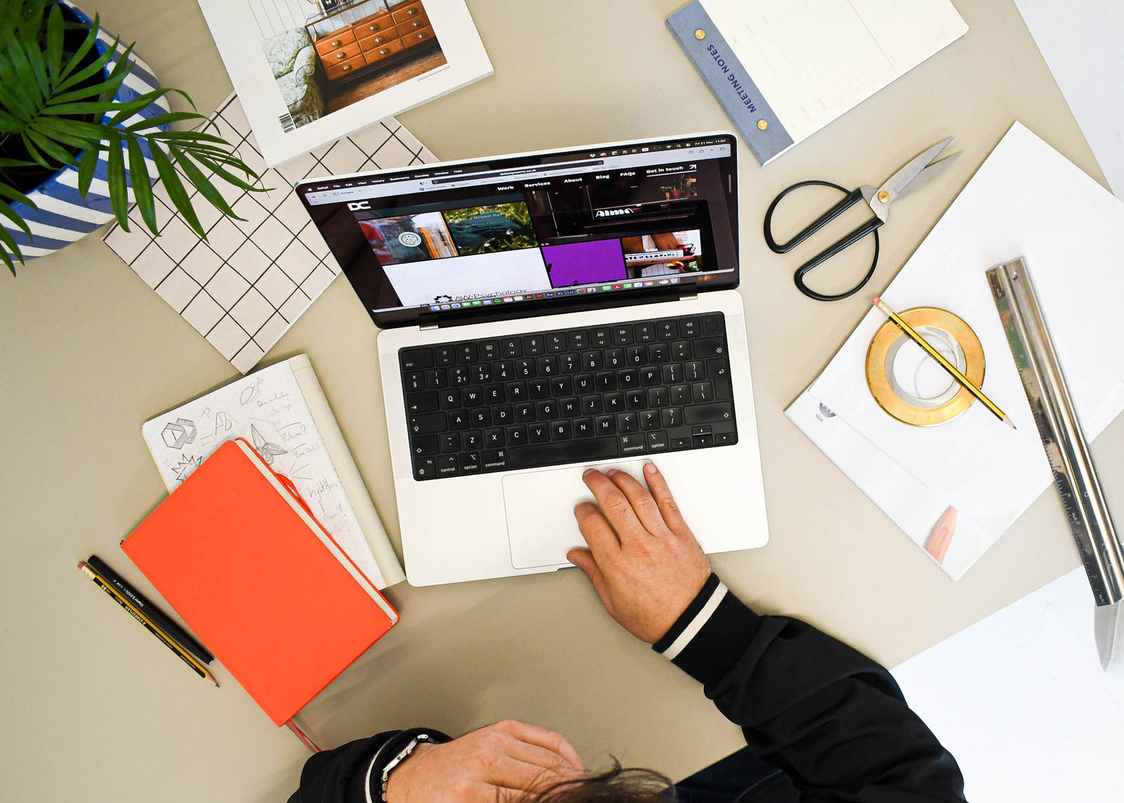 A bird's eye view of Devon branding designer Dennis Cook's desk featuring notebooks, laptop, scissors, and a small pot plant.
