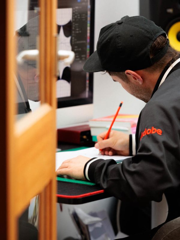 Senior Graphic Designer Dennis Cook works in his Exeter graphic design agency space. Framed by a an open door frame he sits at his desk sketching ideas wearing a black cap, and a black Adobe jacket with a colourful design.
