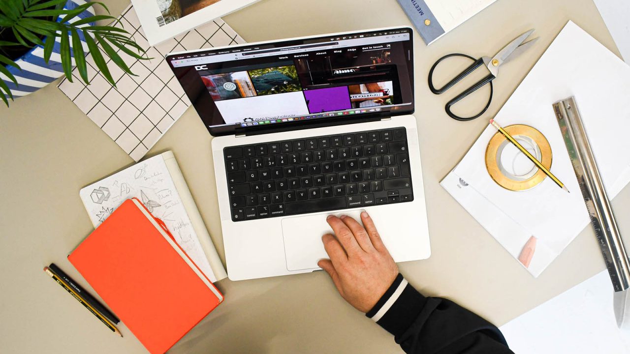 A bird's eye view of Devon branding designer Dennis Cook's desk featuring notebooks, laptop, scissors, and a small pot plant.